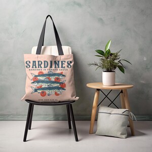 May include: A light pink tote bag with black handles featuring a vintage-style illustration of sardines in tomato sauce. The design includes the word "SARDINES" in blue, along with images of fish and tomatoes. The bag is sitting on a black chair.