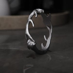 May include: A silver-colored ring with a unique antler design. The ring features a textured surface and a naturalistic, organic shape, with multiple points and curves. The ring is displayed on a dark, reflective surface.