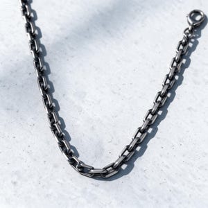 May include: A dark silver-toned chain necklace featuring elongated rectangular links with rounded edges. The metal has a matte, oxidized finish, and the chain is secured with a circular spring ring clasp at one end, creating a uniform link pattern.