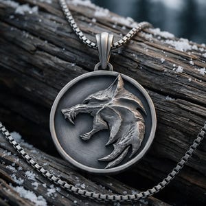 May include: A circular silver-toned metal pendant featuring a relief sculpture of a snarling wolf head in profile. The pendant is suspended from a matching silver-toned box chain necklace with a polished finish and dark recessed detailing.