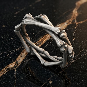 May include: A silver-toned ring crafted from a series of interconnected bone-shaped segments. The metal has a matte, weathered finish with visible joints and anatomical contours forming a continuous circular band structure.