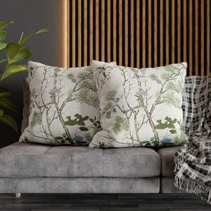 May include: Two decorative throw pillows with a botanical print featuring trees, leaves, and flowers in shades of green, blue, and pink on a cream background. The pillows are placed on a gray sofa.