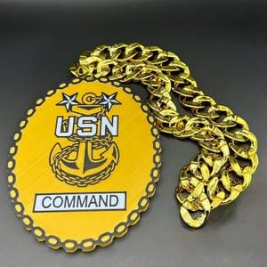 May include: A gold-colored chain and a yellow oval-shaped emblem with the text "USN COMMAND" in white. The emblem features a black anchor and stars. The chain is thick and has a shiny finish.