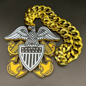May include: A gold chain necklace with a pendant featuring a white eagle with a shield and stars. The shield has a striped design and the eagle is perched on top of an anchor.