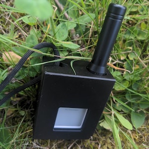 Könnte beinhalten: Ein schwarzes elektronisches Gerät mit einem Bildschirm, auf dem "No Messages" steht, liegt im Gras. Das Gerät hat eine schwarze Antenne und ein schwarzes Kabel. Das Gerät ist rechteckig mit einem quadratischen Bildschirm.