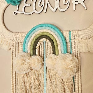 Könnte beinhalten: Makramee-Wandbehang mit Regenbogen-Design in Türkis, Grün und Blau. Das Wort "LEONOR" ist in weißer Schrift dargestellt. Das Stück enthält flauschige Wolkenakzente und türkisfarbene Pompons.