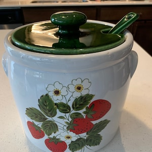 May include: White ceramic jar with a green lid and a spoon. The jar has a strawberry design with red strawberries, green leaves, and white flowers.