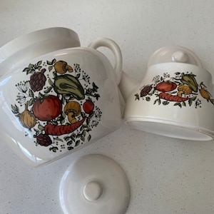 Puede incluir: Una sopera de cerámica blanca con tapa y dos asas. La sopera está decorada con un diseño de verduras coloridas. También hay una tetera de cerámica blanca con tapa y asa. La tetera está decorada con un diseño de verduras coloridas.