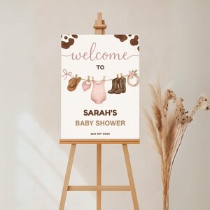 Może przedstawiać: Witamy na Baby Shower Sarah z brązowym tłem w krowie cętki. Na tablicy znajduje się sznurek do suszenia z r&oacute;żowym body dla dziecka, brązowym kowbojskim kapeluszem, r&oacute;żowym śliniakiem i dwiema parami brązowych kowbojskich but&oacute;w.