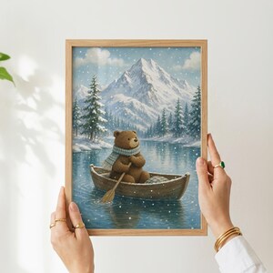 May include: A framed wall art print featuring a brown bear wearing a blue and white striped scarf, sitting in a wooden rowboat on a calm lake. The background shows snow-covered pine trees and a large mountain peak under a light snowfall.
