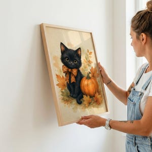 May include: A framed wall art print featuring a black kitten with large eyes wearing an orange polka-dot bow tie, sitting next to a bright orange pumpkin surrounded by autumn leaves in shades of yellow, orange, and brown on a cream background.