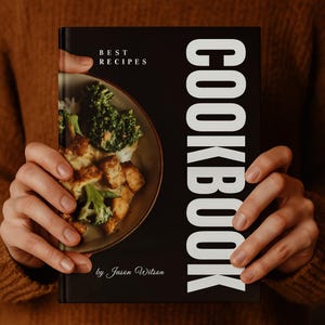Op de afbeelding: Een zwart kookboek dat door een persoon wordt vastgehouden, met de titel "COOKBOOK" in grote witte letters. De omslag toont een foto van een maaltijd met broccoli en kip. De tekst "BEST RECIPES" en de naam van de auteur, "by Jason Wilson", zijn ook zichtbaar.