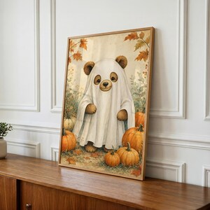 May include: A framed wall art print featuring a brown bear wearing a white ghost costume, standing in a field of orange pumpkins and autumn leaves. The illustration has a soft, warm color palette with detailed textures on the bear and pumpkins.