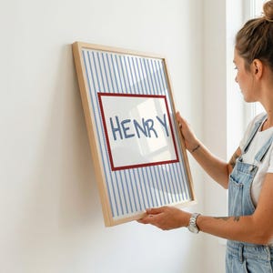May include: A rectangular framed print featuring a light blue and white vertical striped background. In the center, a white rectangular panel with a scalloped red border displays the name HENRY written in a blue, hand-drawn style font.