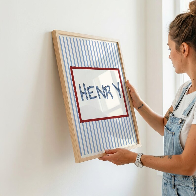 May include: A rectangular framed print featuring a light blue and white vertical striped background. In the center, a white rectangular panel with a scalloped red border displays the name HENRY written in a blue, hand-drawn style font.