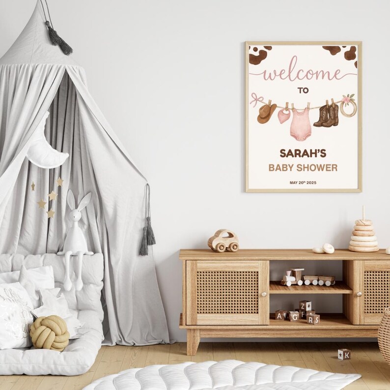 Może przedstawiać: Biały znak z brązowymi akcentami w kształcie krowich plam i tekstem "welcome to Sarah's Baby Shower May 20th, 2023". Znak ma r&oacute;żowy śpioszek dla dziecka, brązowe kowbojskie buty, brązowy kowbojski kapelusz i r&oacute;żową kokardkę wiszącą na sznurku.