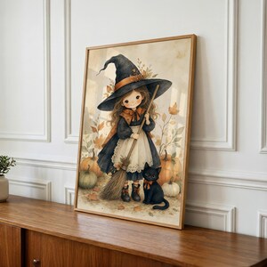 Puede incluir: Una l&aacute;mina de arte enmarcada con una ilustraci&oacute;n en acuarela de un ni&ntilde;o peque&ntilde;o disfrazado de bruja con sombrero y vestido negros, delantal blanco y una escoba. Un gato negro est&aacute; sentado cerca, rodeado de calabazas naranjas y hojas de oto&ntilde;o.