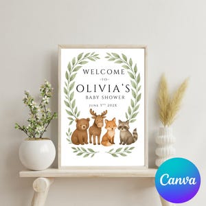May include: A white sign with a green leafy wreath around the text "Welcome to Olivia's Baby Shower June 3rd 202X".  Four woodland animals, a bear, moose, fox, and raccoon, are standing in front of the wreath.