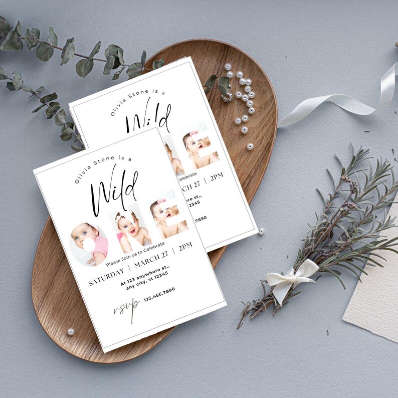 May include: White invitation cards with the text "Olivia Stone is a Wild" and baby photos. The cards are on a wooden tray with decorative elements. The invitation includes the date, time, and RSVP information.