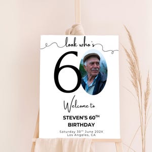 May include: A white welcome sign with a black number 60 and the text "look who's 60". A photo of a man is in the background. The text "Welcome to STEVEN'S 60TH BIRTHDAY" is below the number. The date and location are listed below the birthday text.