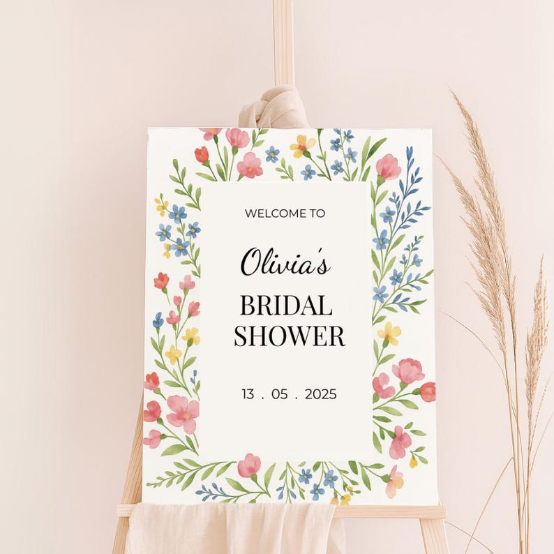 May include: A white welcome sign with a floral border in shades of pink, blue, and yellow. The text reads "Welcome to Olivia's Bridal Shower 13.05.2025".