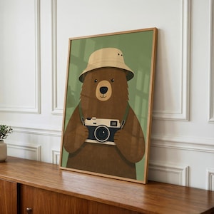 May include: A framed wall art print featuring a brown bear wearing a beige bucket hat. The bear is holding a vintage-style camera with a black and white body. The background of the illustration is a solid muted olive green color.