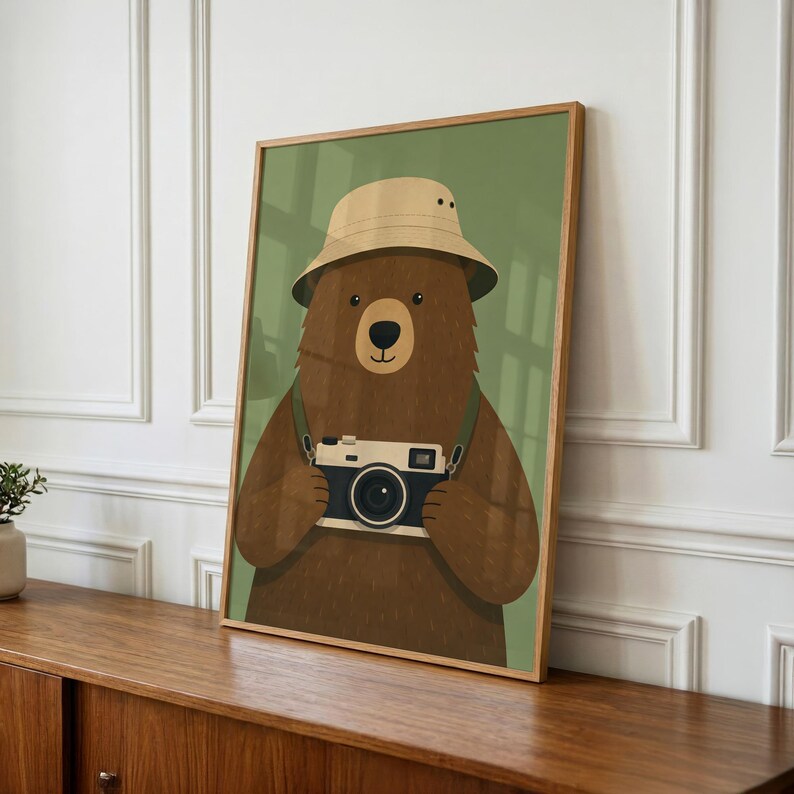 May include: A framed wall art print featuring a brown bear wearing a beige bucket hat. The bear is holding a vintage-style camera with a black and white body. The background of the illustration is a solid muted olive green color.
