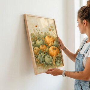 May include: A framed wall art print featuring a watercolor illustration of several orange and green pumpkins nestled among green foliage and small yellow flowers. The artwork is encased in a light-colored wooden frame with a clean, simple border.