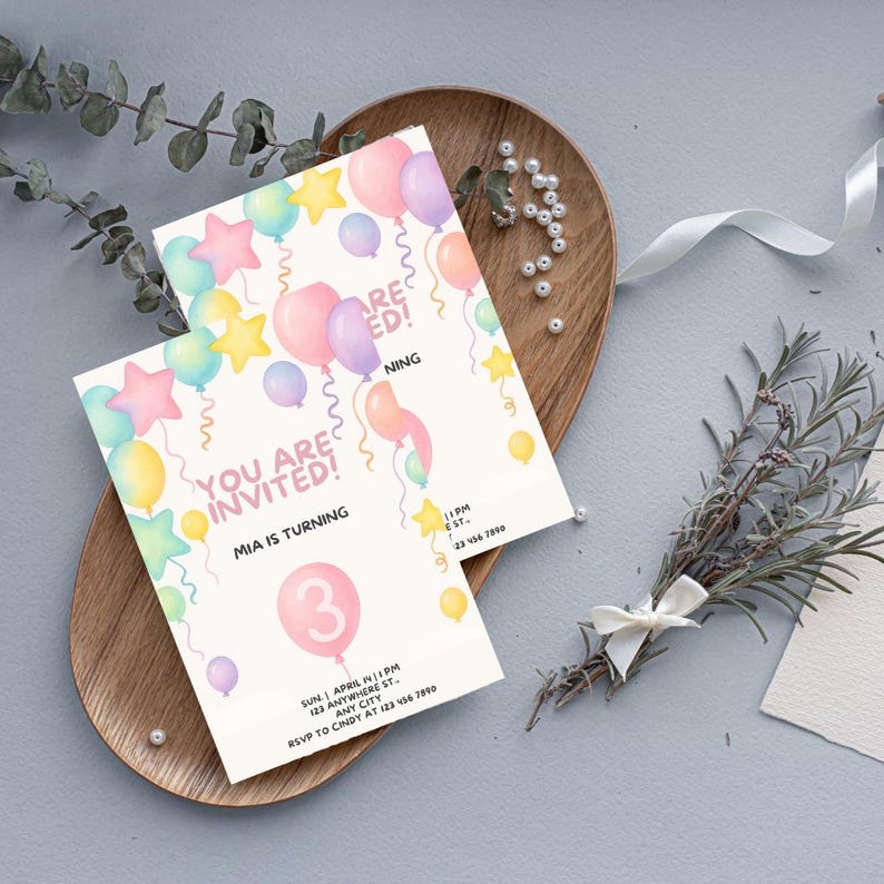 Op de afbeelding: Twee rechthoekige verjaardagsuitnodigingen met pastelkleurige ballonnen en sterren. De tekst luidt YOU ARE INVITED! MIA IS TURNING 3, met tijdelijke aanduidingen voor datum, tijd en locatie. De kaarten liggen op een ovaal houten dienblad.