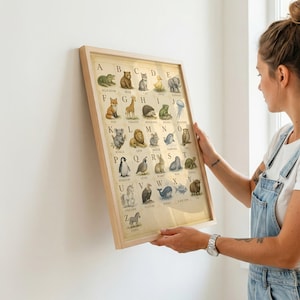 Puede incluir: Un p&oacute;ster del alfabeto enmarcado que presenta una cuadr&iacute;cula de animales ilustrados correspondientes a cada letra de la A a la Z. La impresi&oacute;n tiene un fondo beige claro con letras negras y dibujos detallados de animales en un marco de madera clara.