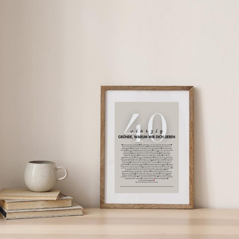 Puede incluir: Un marco con el texto en alem&aacute;n "40 Gr&uuml;nde, Warum Wir Dich Lieben". La impresi&oacute;n tiene un fondo gris claro y un marco de madera. Libros y una taza est&aacute;n sobre una superficie de madera.