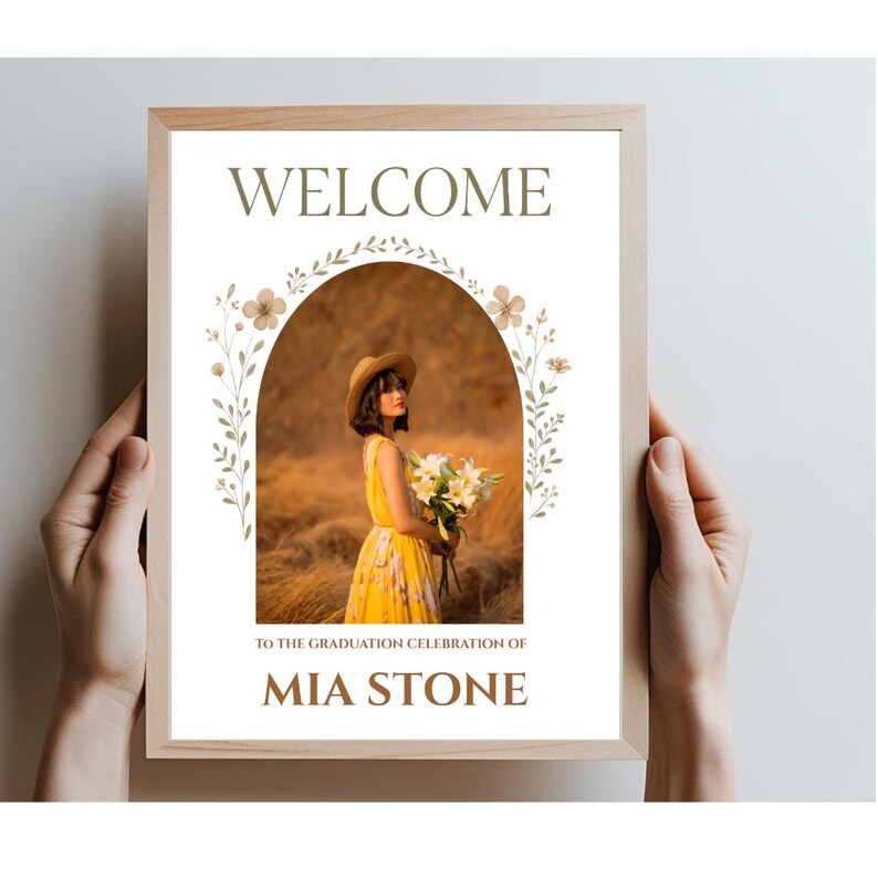 May include: Welcome to the graduation celebration of Mia Stone. A framed sign with a floral archway and a photo of a woman in a yellow dress holding a bouquet of white flowers.