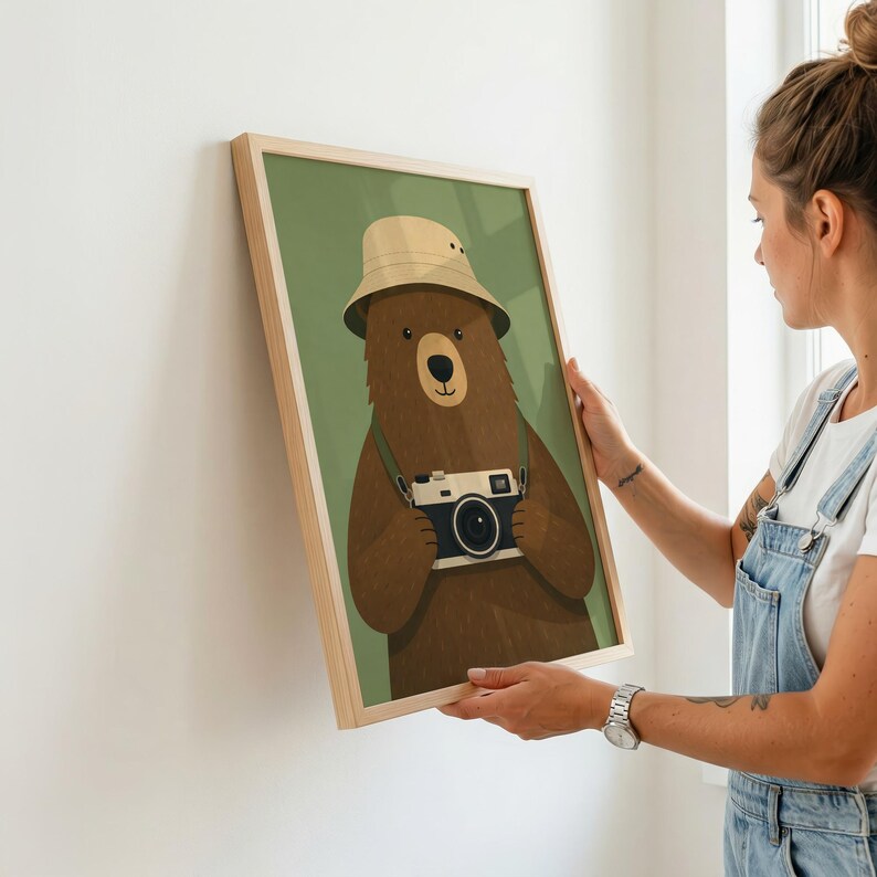 May include: A framed wall art print featuring a brown bear wearing a beige bucket hat and holding a vintage-style camera. The bear is centered against a solid olive green background, and the artwork is enclosed in a light-colored wooden frame.