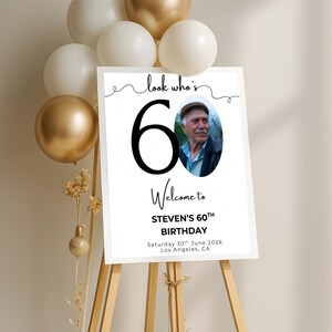May include: A white welcome sign with black text and a photo of a man. The sign says "look who's 60", "Welcome to STEVEN'S 60TH BIRTHDAY", "Saturday 30th June 202X", and "Los Angeles, CA".
