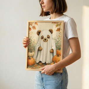 May include: A rectangular framed print featuring an illustration of a brown bear wearing a white ghost costume. The bear is surrounded by orange pumpkins and autumn leaves in a soft, muted color palette. The artwork is set against a light background.