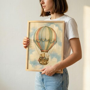 Może przedstawiać: Oprawiony wydruk akwarelowy przedstawiający duży, pasiasty balon na ogrzane powietrze w delikatnych pastelowych odcieniach błękitu, r&oacute;żu i kremu. Mały pluszowy miś i kr&oacute;lik siedzą w wiklinowym koszu zawieszonym pod balonem na tle zachmurzonego nieba.
