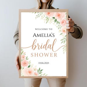 Op de afbeelding: Een wit bord met een bloemenontwerp in roze en groene tinten. Op het bord staat "Welkom bij Amelia's bruidsdouche 03.06.2025".