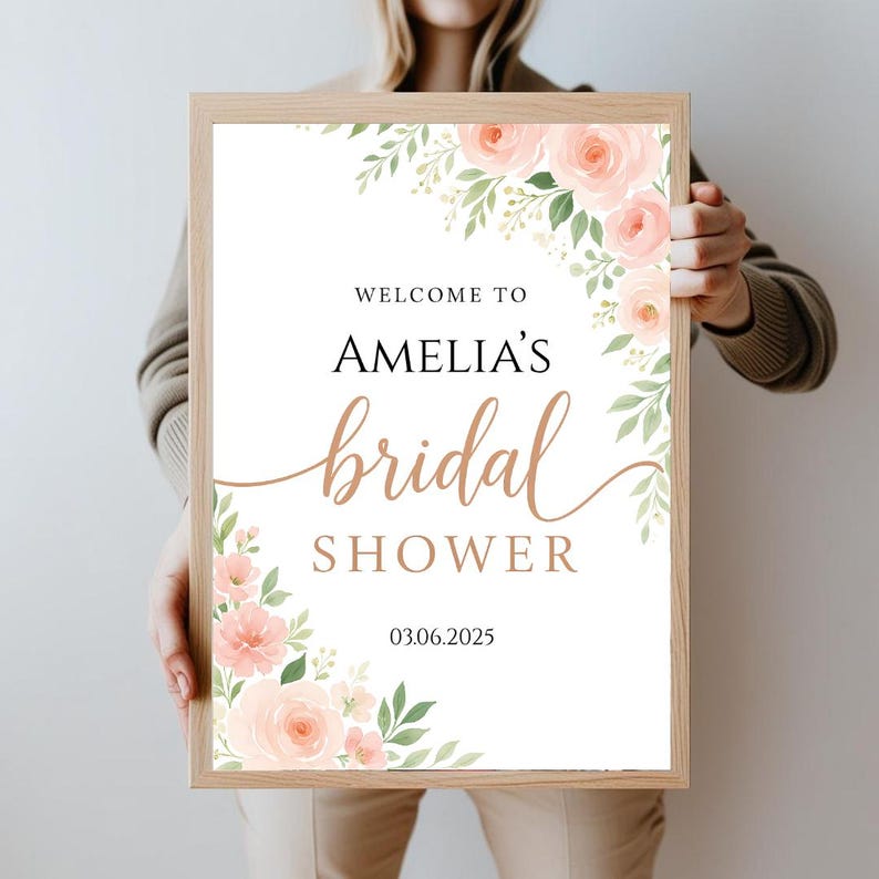 Op de afbeelding: Een wit bord met een bloemenontwerp in roze en groene tinten. Op het bord staat "Welkom bij Amelia's bruidsdouche 03.06.2025".