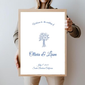 May include: A framed wedding welcome sign with a white background and blue text. The sign reads "Welcome to the wedding of Olivia & Liam" with the date July 1st, 2025, and Santa Barbara, California. A floral bouquet illustration is centered.