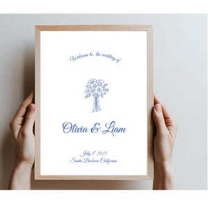 May include: A framed wedding sign with a light wood frame. The white background features blue script that reads "Welcome to the wedding of Olivia & Liam" with a floral bouquet illustration. The date is July 1st, 2025, in Santa Barbara, California.
