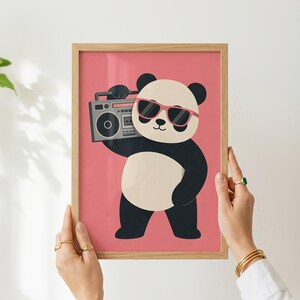 May include: A framed wall art print featuring a cartoon panda with black and white fur, wearing pink-rimmed sunglasses, and holding a grey boombox. The background of the illustration is a solid pink color, and the frame is light-colored wood.