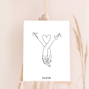 May include: A black and white line art print of two hands holding each other with a heart in the middle. The names "Anna" and "Casey" are written on either side of the heart. The text "You & Me" is written below the hands.