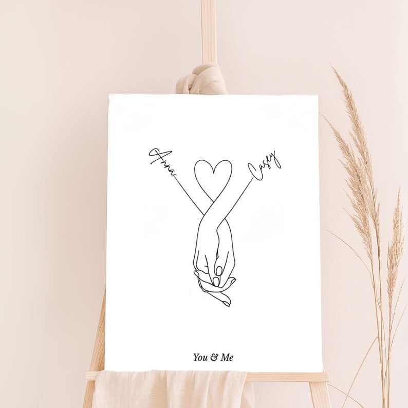 May include: A black and white line art print of two hands holding each other with a heart in the middle. The names "Anna" and "Casey" are written on either side of the heart. The text "You & Me" is written below the hands.