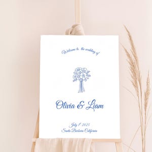 May include: A white wedding welcome sign on an easel, featuring blue text and a floral illustration. The sign reads "Welcome to the wedding of Olivia & Liam" with the date July 1st, 2025, and location Santa Barbara, California.