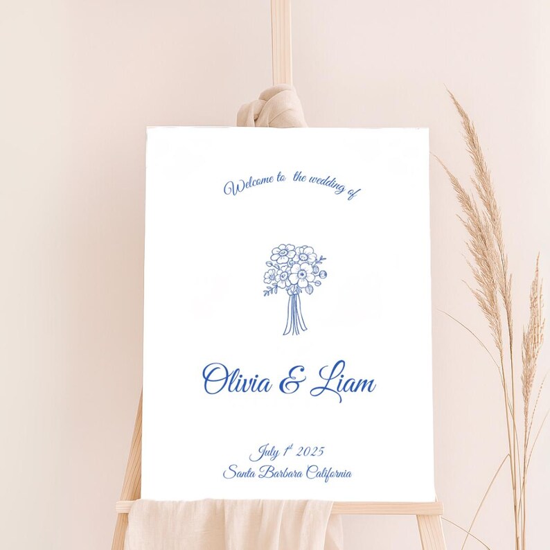 May include: A white wedding welcome sign on an easel, featuring blue text and a floral illustration. The sign reads "Welcome to the wedding of Olivia & Liam" with the date July 1st, 2025, and location Santa Barbara, California.
