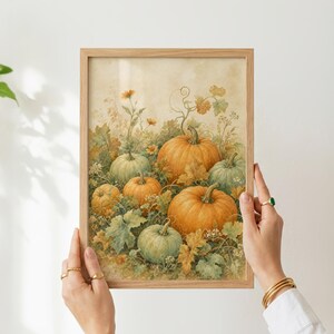 May include: A framed watercolor-style print featuring a harvest scene with multiple orange and sage green pumpkins nestled among sprawling vines, broad leaves, and small wildflowers against a soft, neutral, cream-colored background.