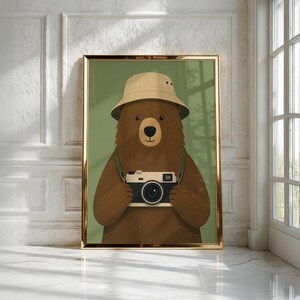 May include: A framed wall art print featuring a brown bear wearing a beige bucket hat and holding a vintage-style camera. The bear is centered against a solid olive green background, and the artwork is enclosed in a thin metallic gold frame.