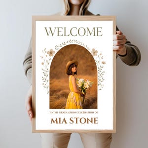 May include: A welcome sign with a floral archway and a photo of a person in a yellow dress and a straw hat. The text on the sign reads "Welcome to the graduation celebration of Mia Stone".