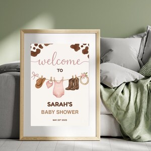 Może przedstawiać: Witamy na Baby Shower Sarah z brązowym tłem w kropki krowy. Na plakacie znajduje się sznurek do suszenia z r&oacute;żowym body dla niemowlęcia, r&oacute;żowym śliniakiem, brązowym kowbojskim kapeluszem i dwiema parami brązowych kowbojskich but&oacute;w.