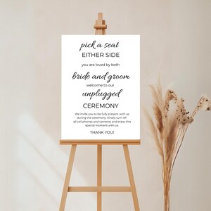 May include: White sign with black text that reads "pick a seat either side you are loved by both bride and groom welcome to our unplugged ceremony we invite you to be fully present with us during the ceremony kindly turn off all cell phones and cameras and enjoy this special moment with us thank you!"